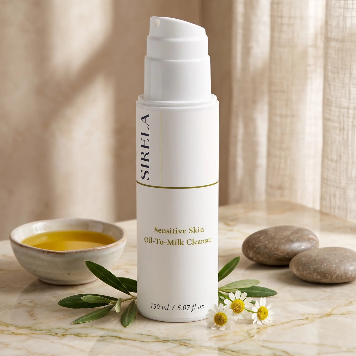 Sirela Sensitive Skin Oil-To-Milk Cleanser bottle on marble beside a ceramic bowl of olive oil, olive branch, chamomile flowers and smooth river stones