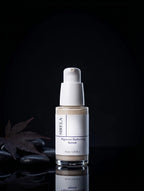 Sirela Pigment Perfecting Serum in a clear glass bottle with a white pump dispenser, set against a dark background with decorative stones and a leaf.