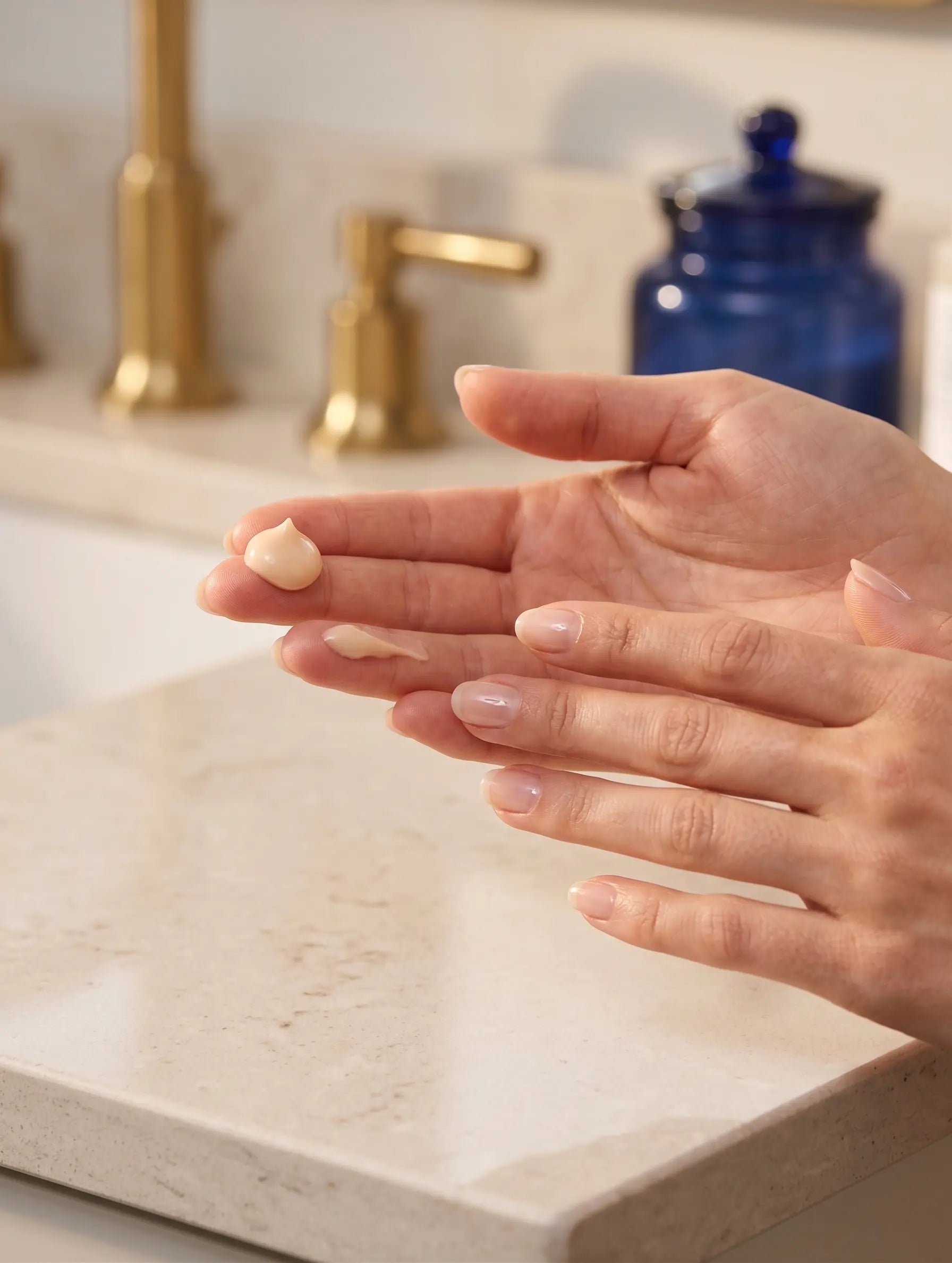 A close-up of a person's hands applying or spreading a small dollop of cream or lotion on the back of one hand, set in a bright bathroom with a marble countertop, gold faucet, and a blue glass jar in the background.