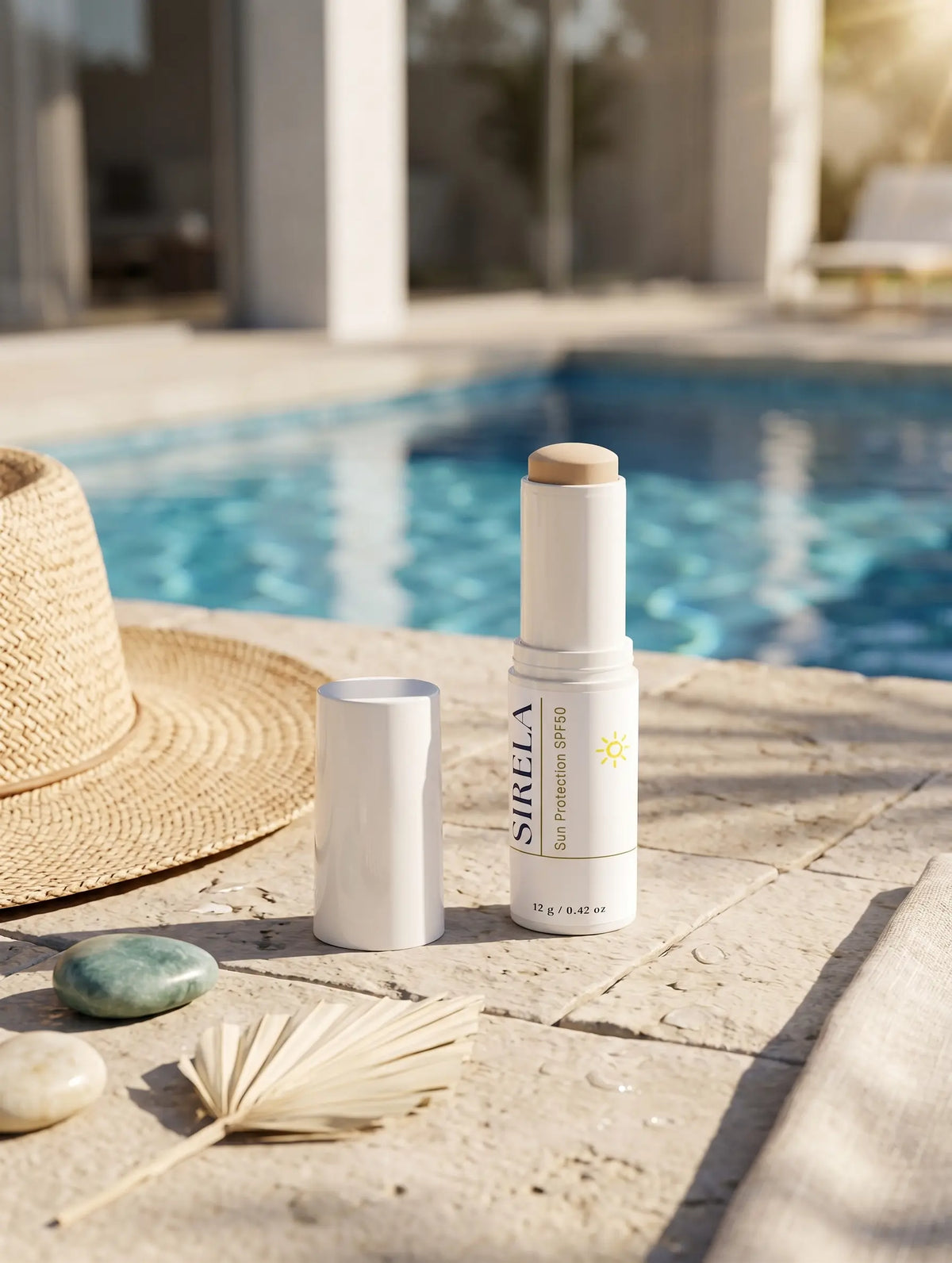 A white stick sunscreen bottle labeled SIRELA SPF 50+ with its cap removed, placed on a stone patio next to a straw hat, stones, and a dried palm leaf with a swimming pool in the background.